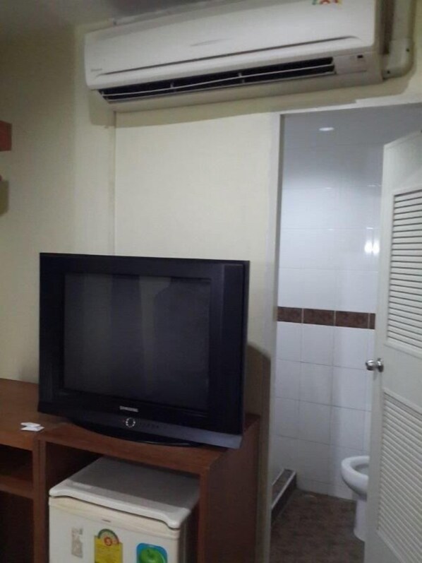 Una habitación con una televisión sobre un soporte de madera, un aire acondicionado arriba, y un baño con inodoro y puerta.