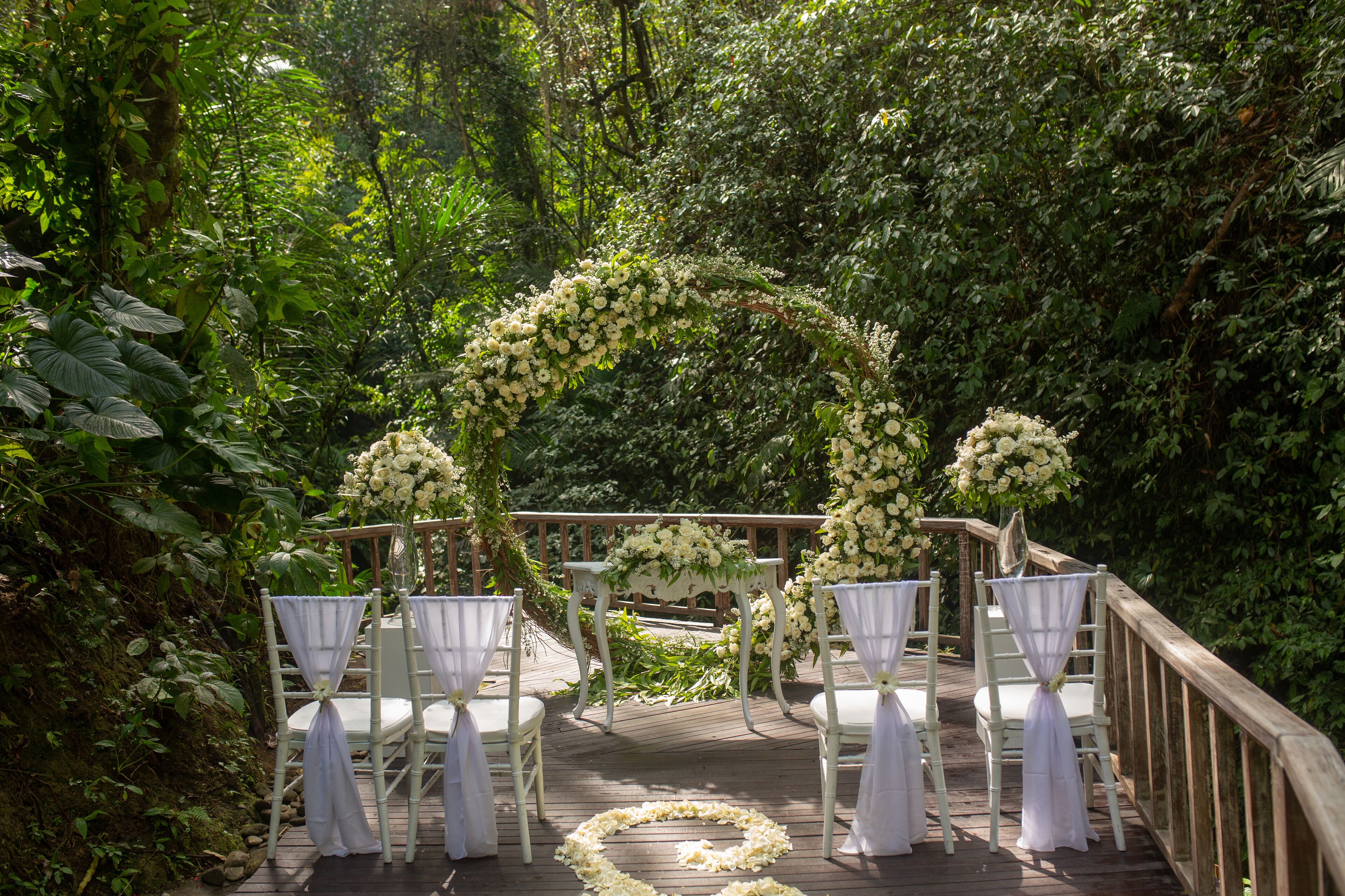 Una terraza de madera con sillas blancas y un arco floral, rodeada de abundante vegetación.