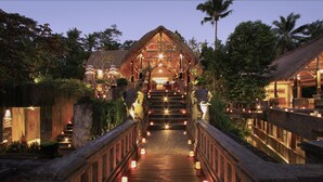 A wooden bridge with steps leading to a thatched-roof structure, surrounded by greenery and palm trees. - The Kayon Resort (Ubud)