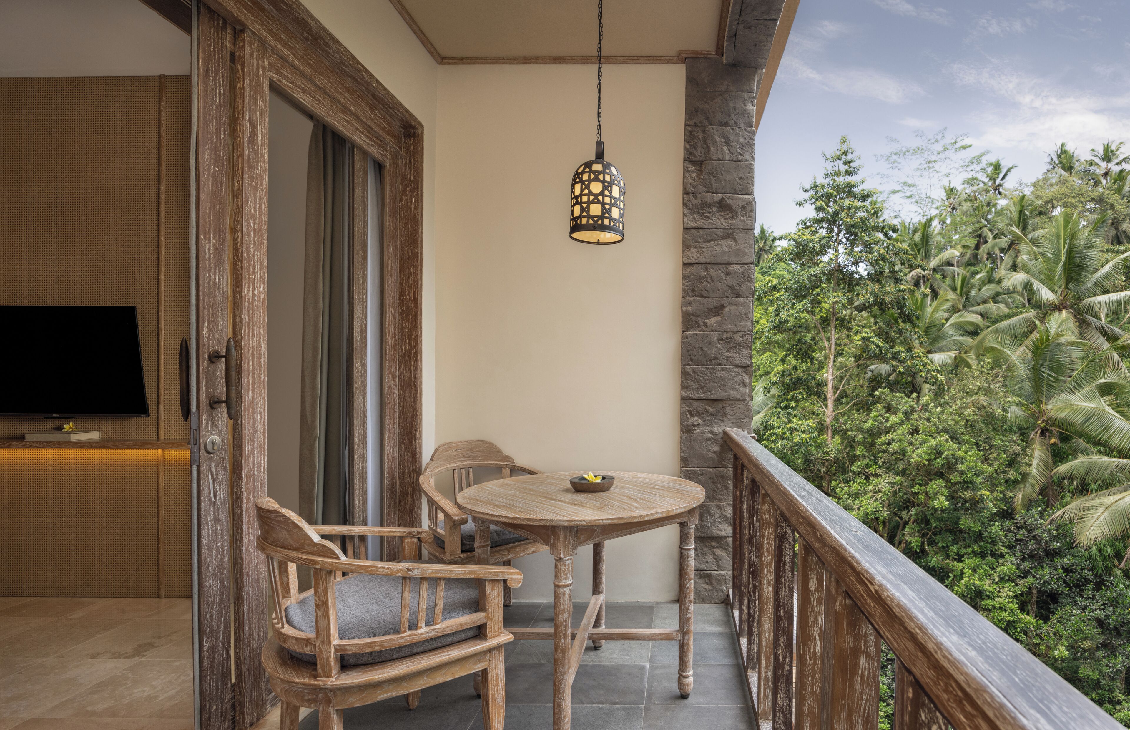 A wooden balcony with a table and chairs, a hanging light fixture, and a view of tropical vegetation.