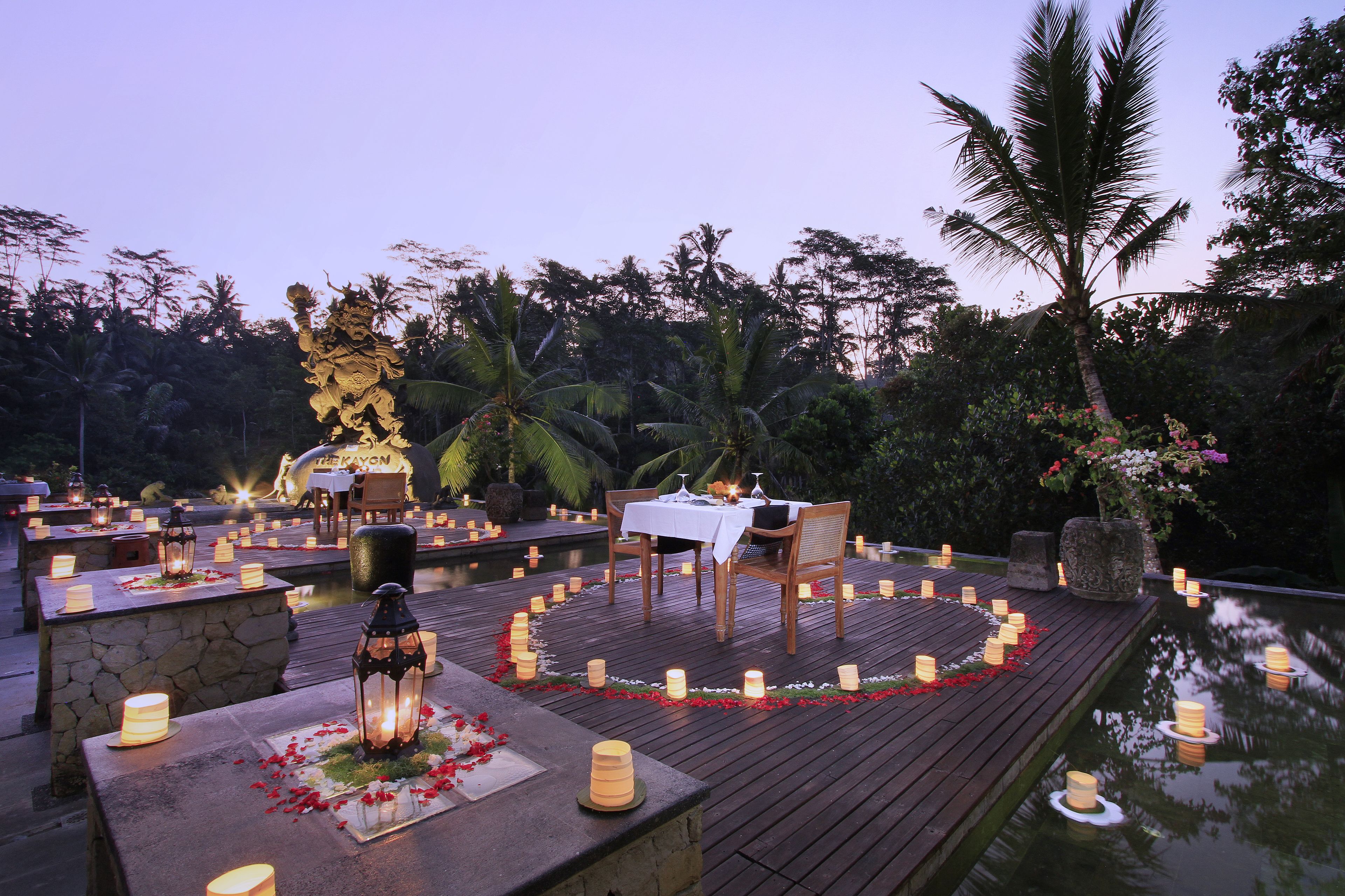 An outdoor dining area with tables set for two, surrounded by a circular arrangement of candles and flower petals, overlooking a body of water.