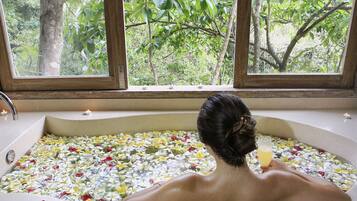 A bathtub filled with water and flower petals, with a view of trees through a window.
