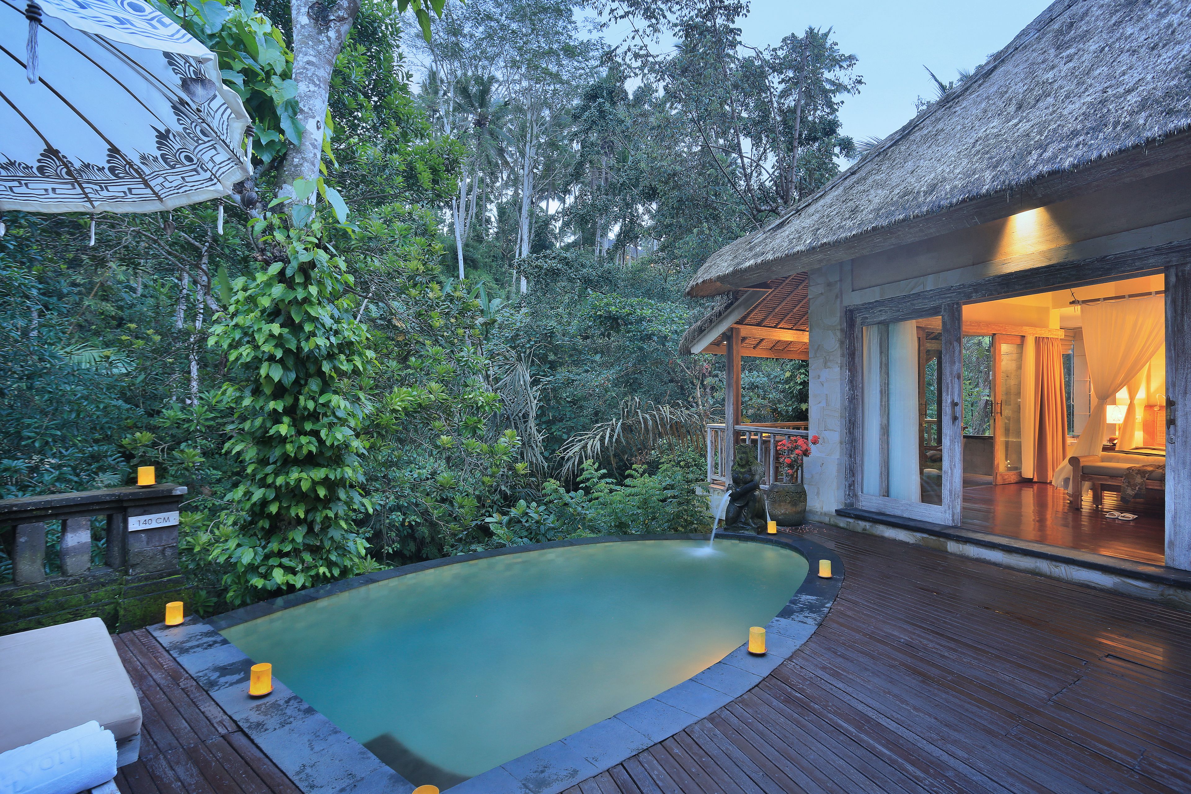A wooden deck with a pool, surrounded by lush greenery and a thatched-roof structure.