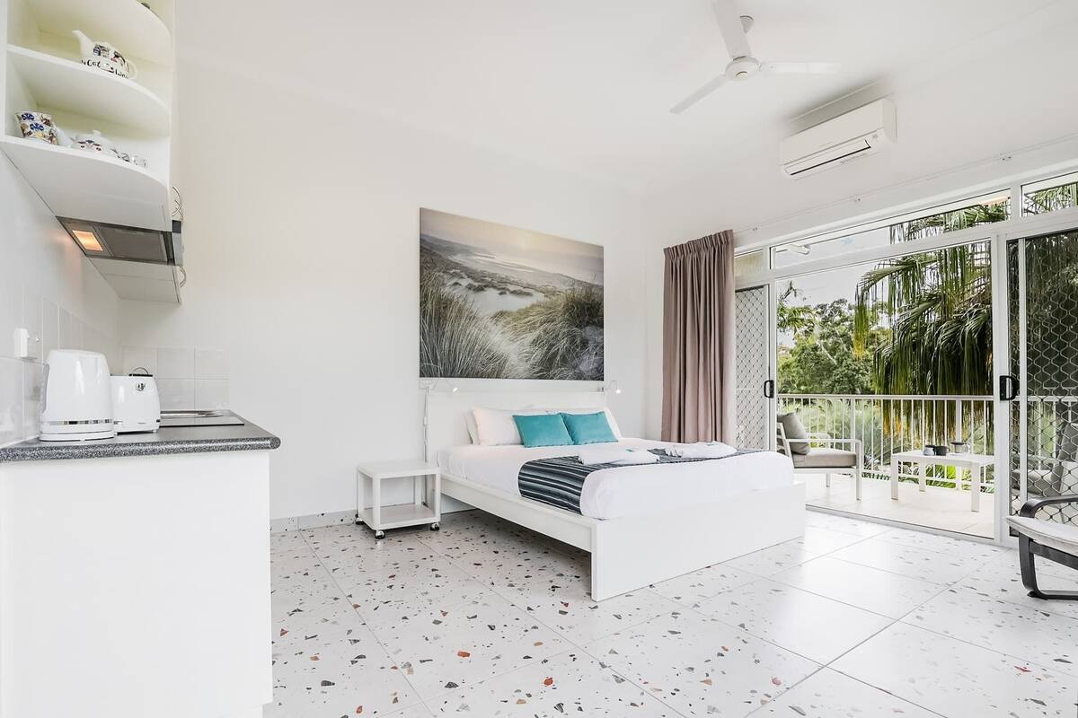 A modern hotel room with a bed, a small bedside table, a coffee maker, and a large window with a view of greenery.
