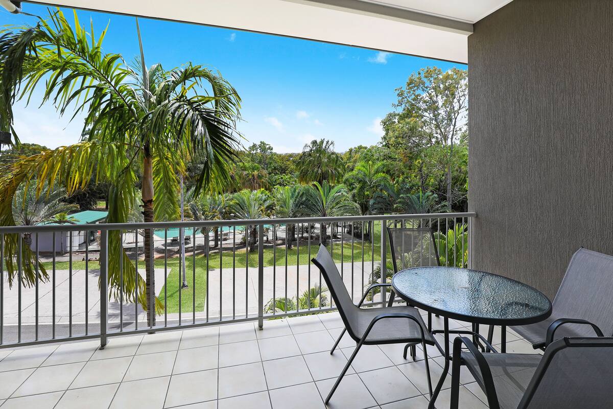 A balcony with a glass table and chairs, a view of a green lawn, and a palm tree.
