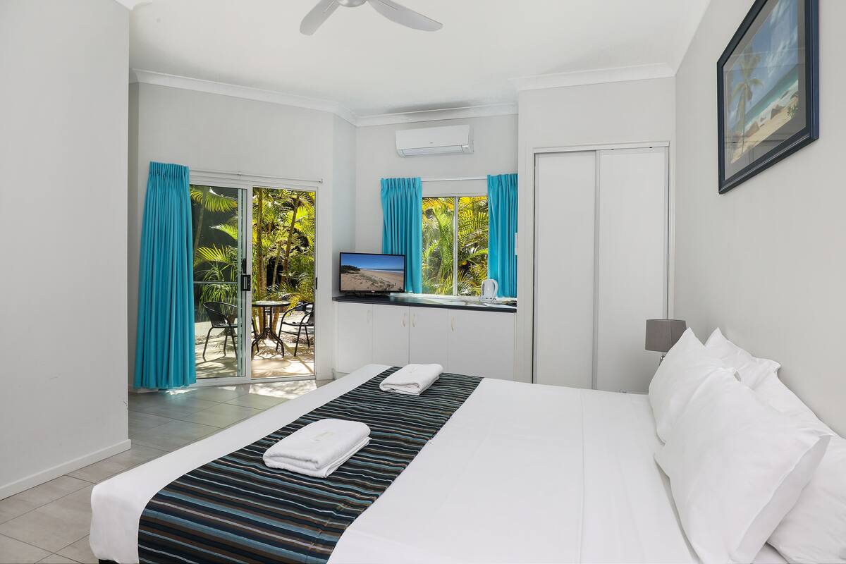 A hotel room with a bed, a desk with a computer, a window with curtains, and a view of greenery outside.
