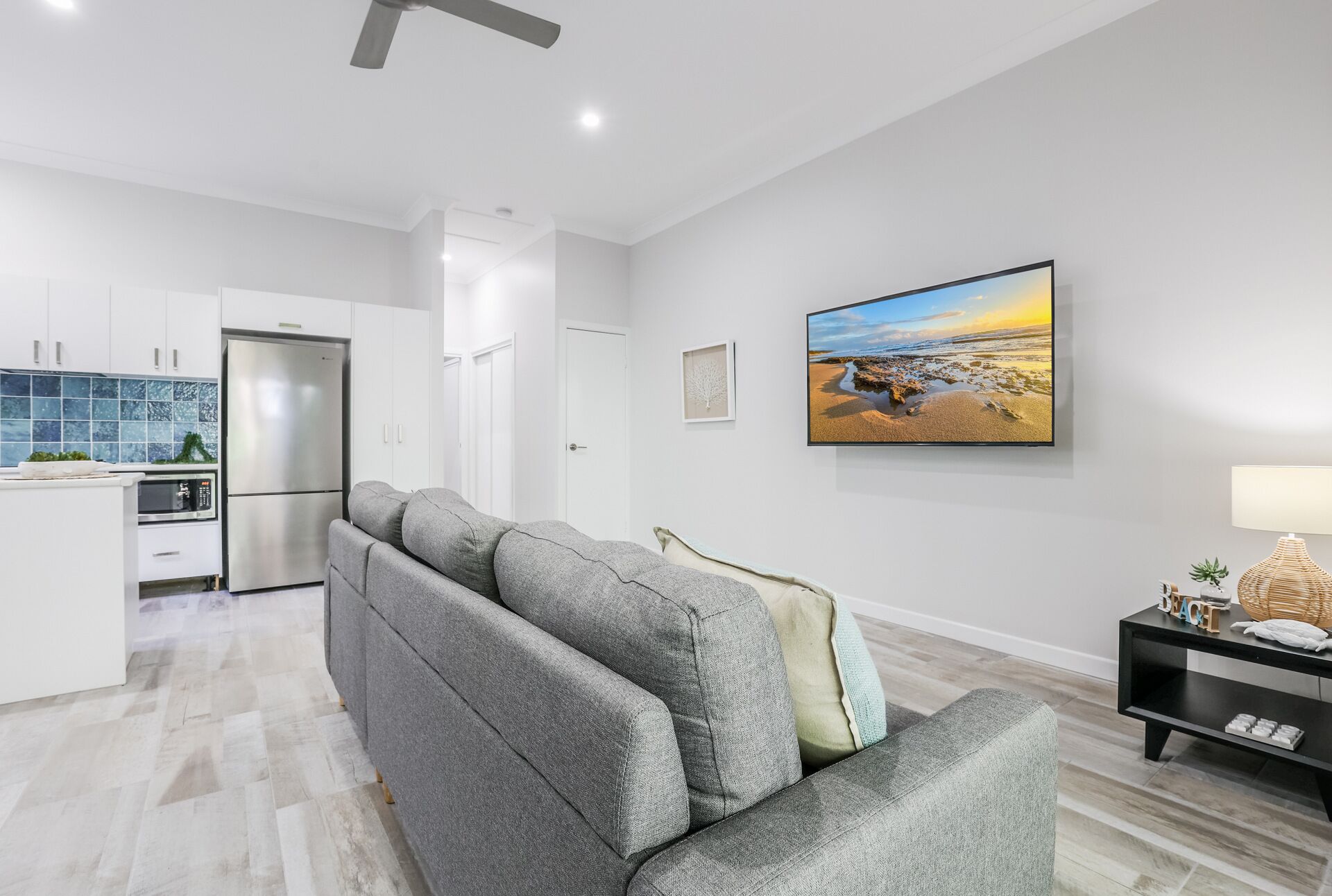 A modern living room with a grey sofa, a flat-screen TV, and a kitchen in the background.