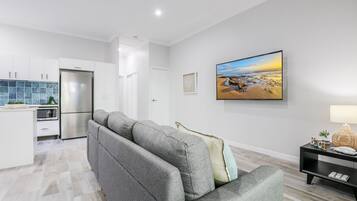 A modern living room with a grey sofa, a flat-screen TV, and a kitchen in the background.
