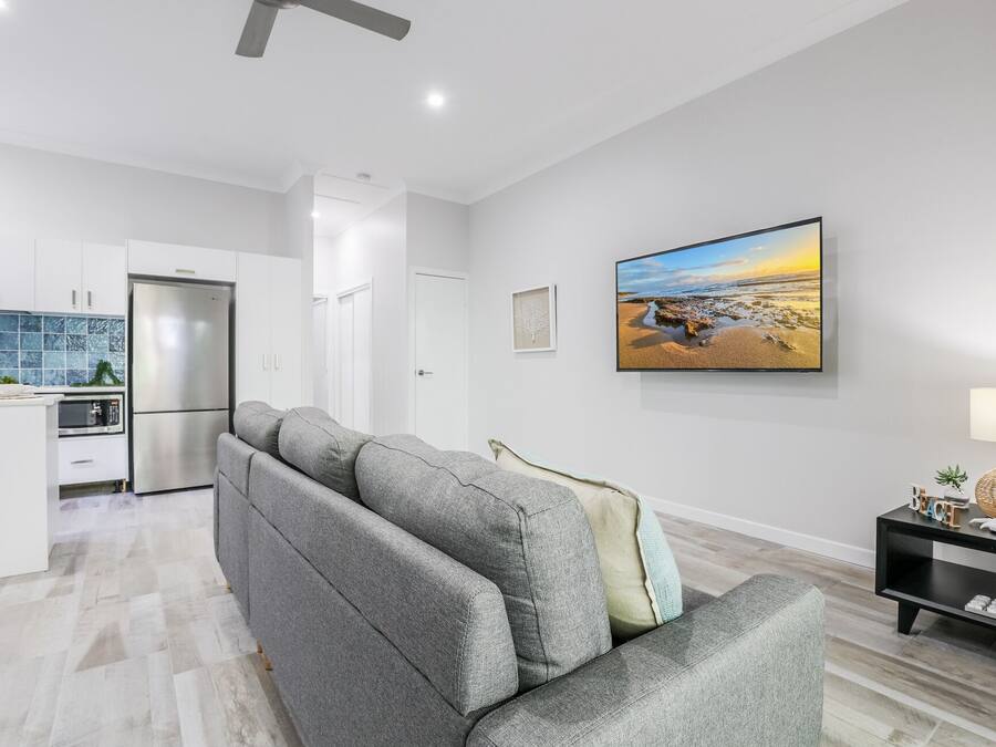 A modern living room with a grey sofa, a flat-screen TV, and a kitchen in the background.