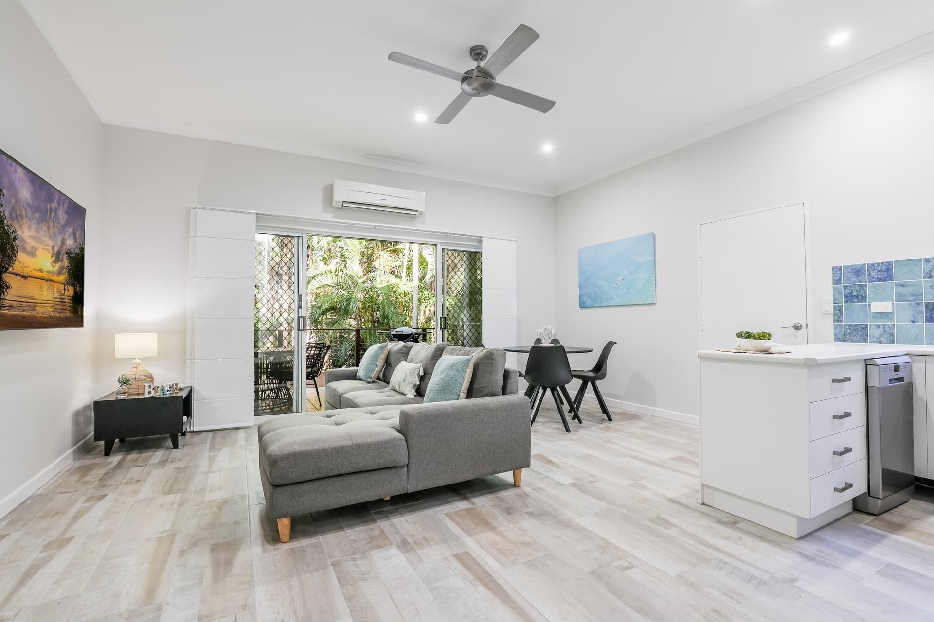 Un salon moderne avec un canapé gris, une table à manger avec des chaises, un ventilateur de plafond et un coin cuisine équipé d’un lave-vaisselle.
