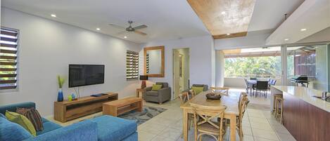 A modern living room with a flat-screen TV, blue sofas, a wooden dining table, and a kitchen area with a grill.
