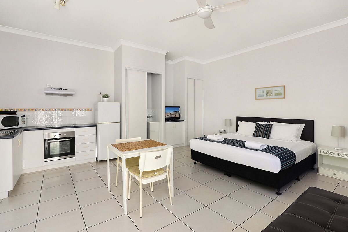 A room with a bed, a dining table with chairs, a kitchen area, and a ceiling fan.