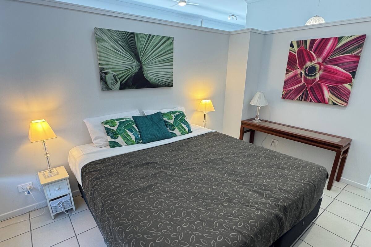 A neatly arranged bedroom with a bed, two bedside lamps, a nightstand, a wooden bench, and two wall-mounted artworks.