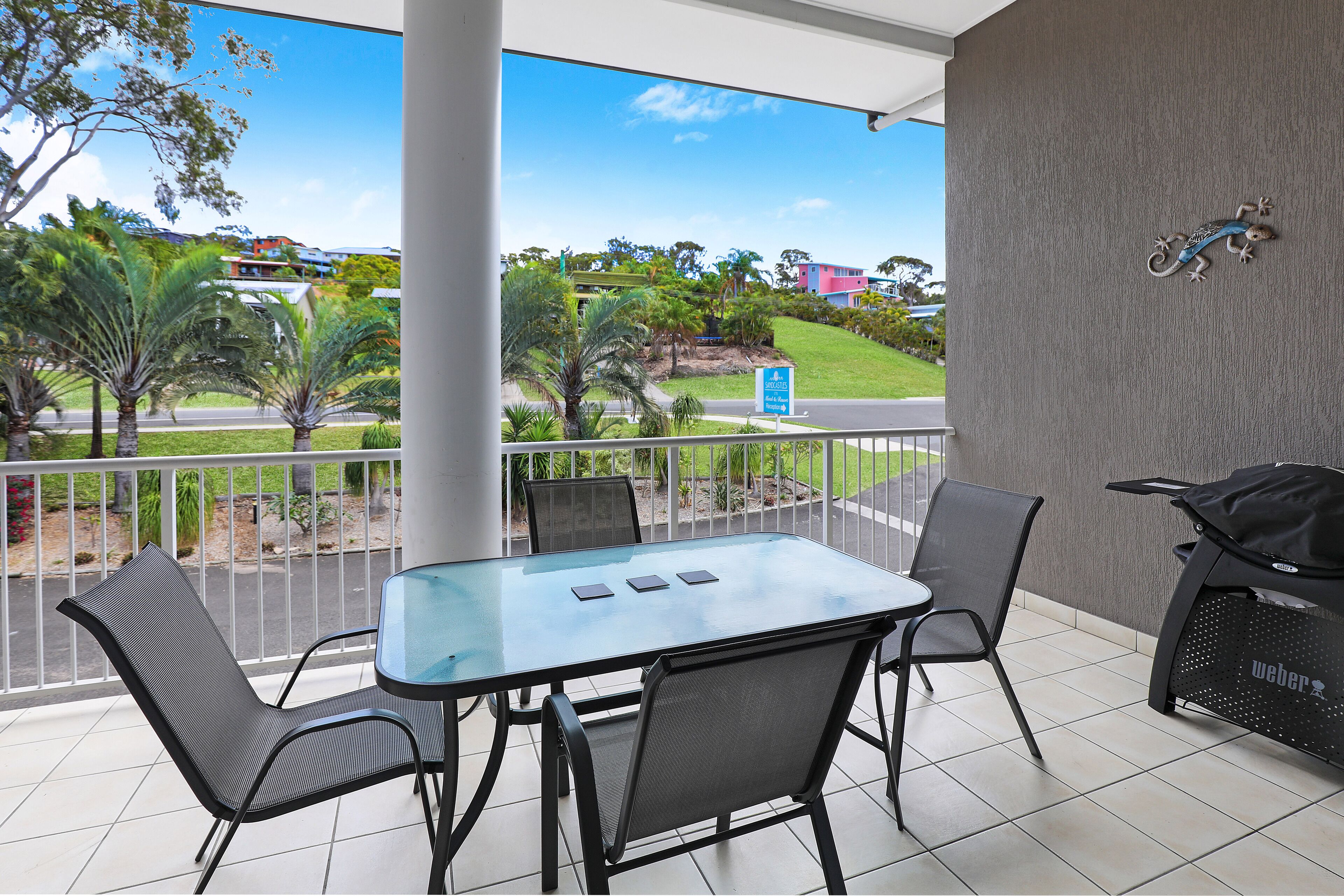 A balcony with a glass table and chairs, a grill, and a view of a road and greenery.