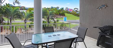 A balcony with a glass table and chairs, a grill, and a view of a road and greenery.