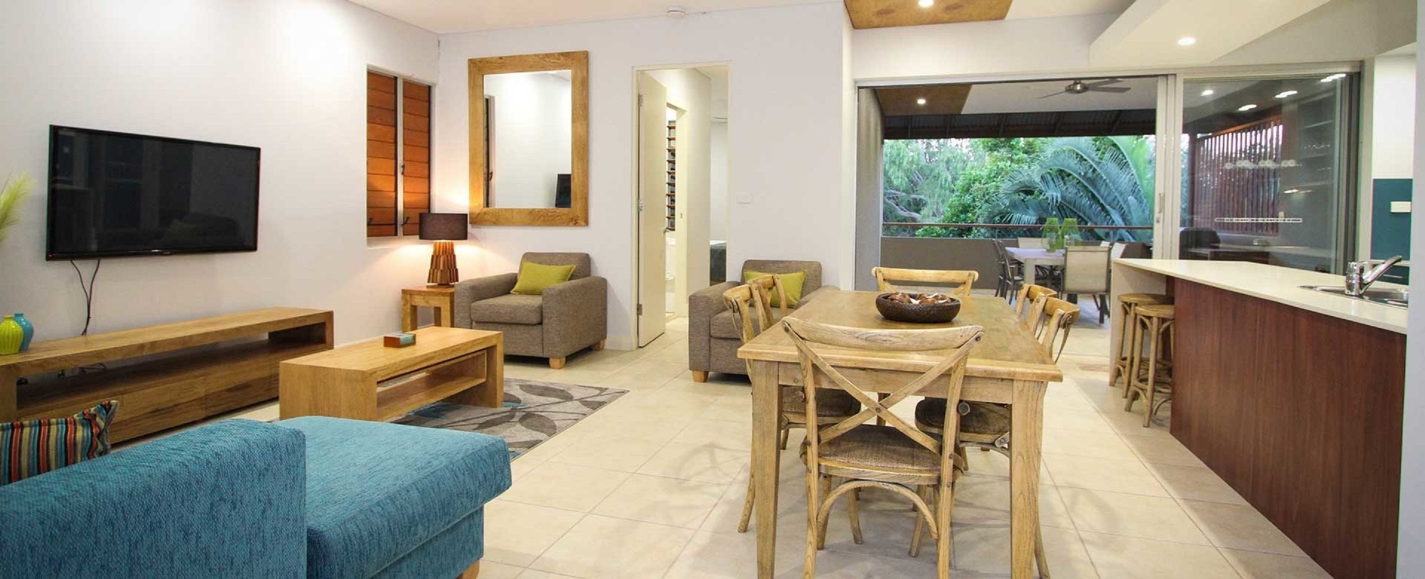 A modern living room with a flat-screen TV, a sofa, a coffee table, and a dining area with wooden chairs and a table.