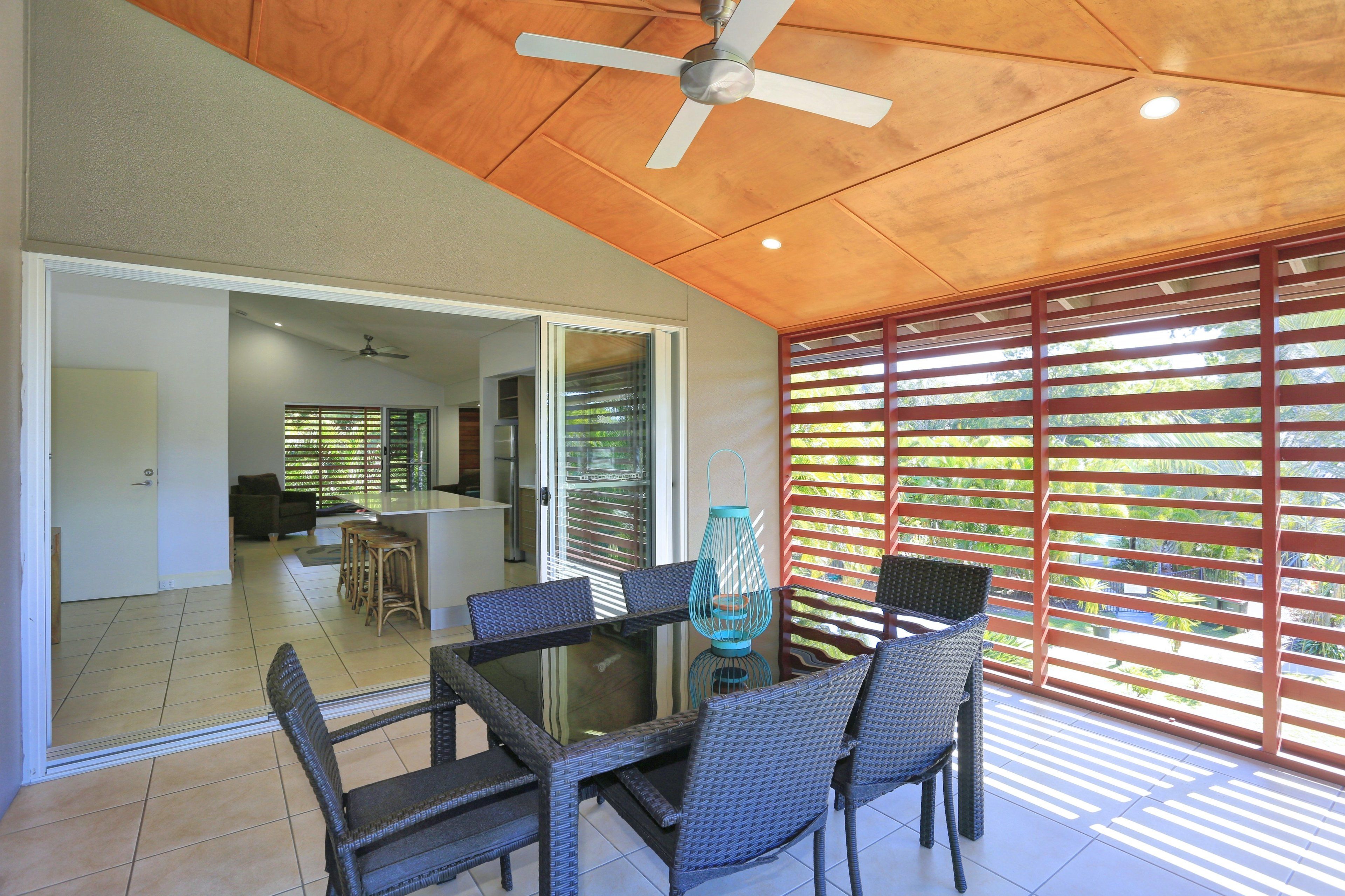 Uma sala de jantar com mesa de vidro e cadeiras de vime, um ventilador de teto e vista para uma cozinha e área de estar através de uma porta deslizante.