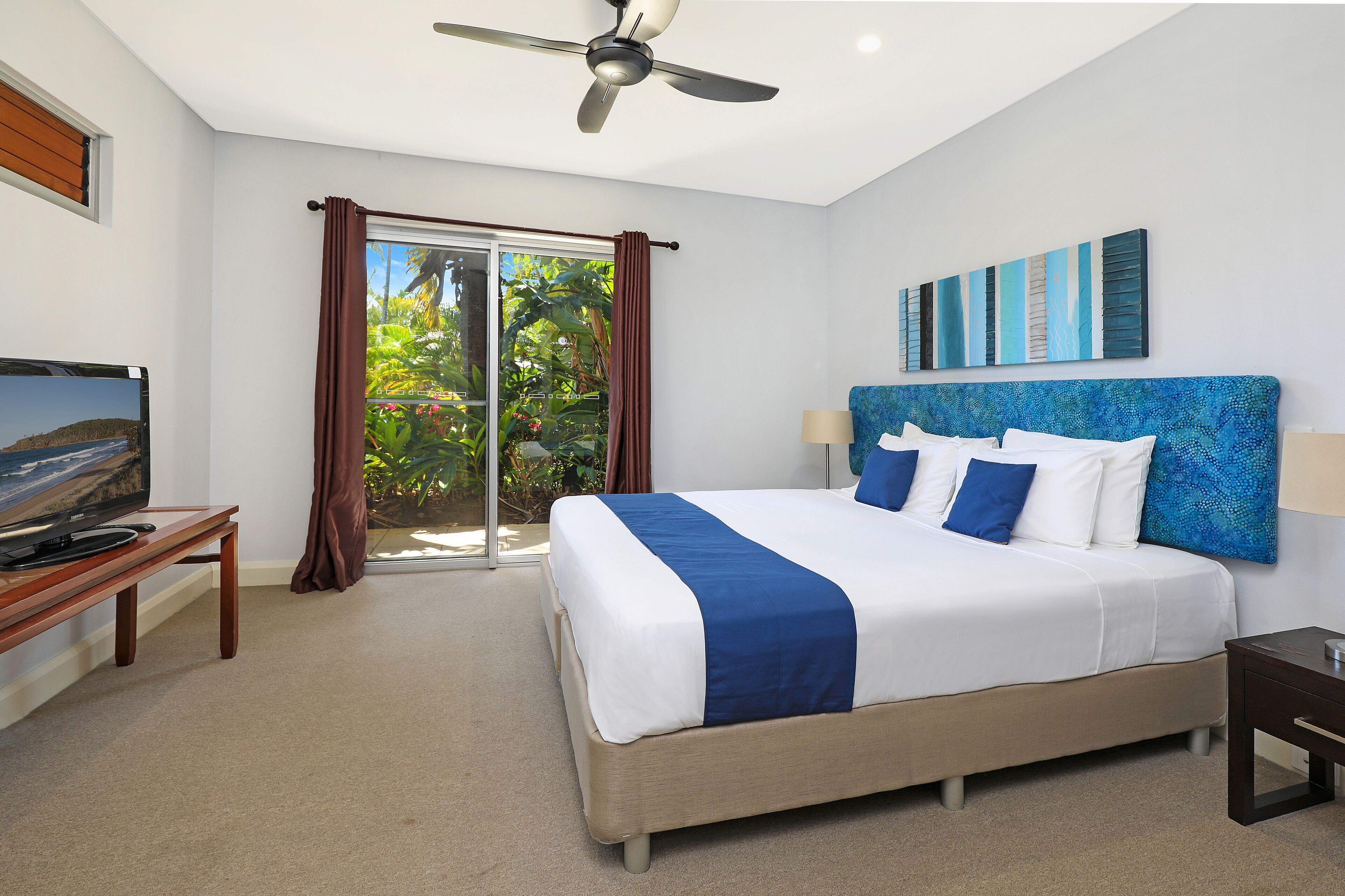 Une chambre d’hôtel équipée d’un lit, d’une télévision, d’un ventilateur de plafond, d’une fenêtre avec des rideaux et offrant une vue sur la verdure environnante.