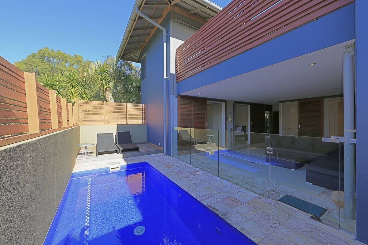 A modern outdoor pool area with a glass enclosure, lounge chairs, and a wooden deck.