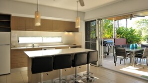 Cozinha moderna com ilha central, banquetas de bar e porta de correr de vidro que dá para área externa com churrasqueira.