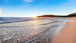 Uma praia com ondas suaves batendo na costa, céu claro e uma colina ao longe.