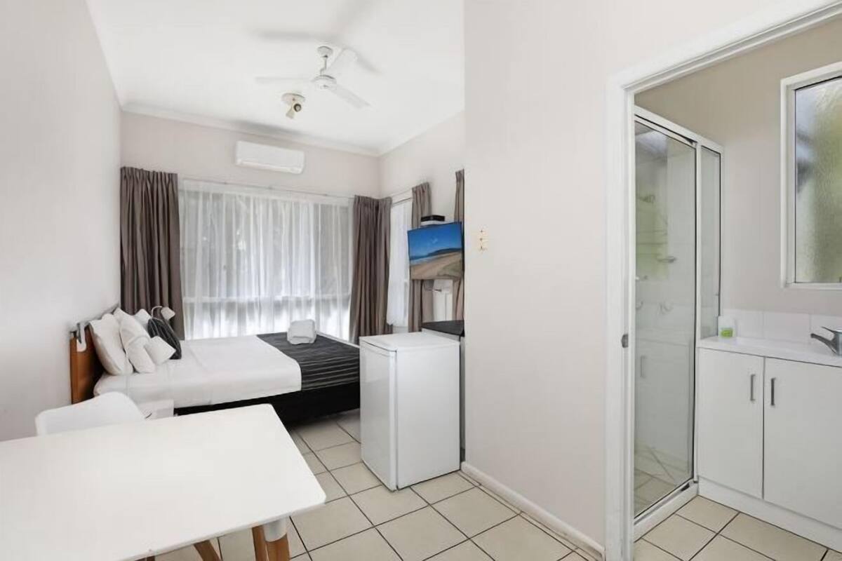 A compact hotel room with a bed, a small desk, a refrigerator, a shower area, and a ceiling fan.