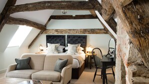 A cozy attic bedroom with exposed wooden beams, a large bed with multiple pillows, a sofa, a table, and a chair. - Kings Head (Cirencester)
