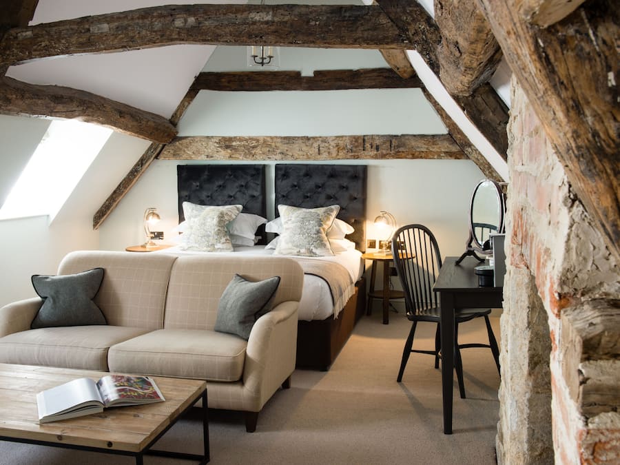 A cozy attic bedroom with exposed wooden beams, a large bed with multiple pillows, a sofa, a table, and a chair.