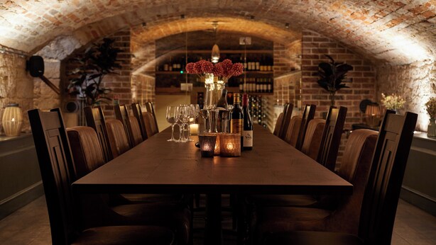 A long wooden dining table with chairs, a vase with flowers, and wine bottles in a brick-walled room with a vaulted ceiling. - Kings Head (Cirencester)