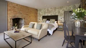 A room with a brick fireplace, a bed with a headboard, a sofa, a dining table with chairs, and a vase of flowers. - Kings Head (Cirencester)
