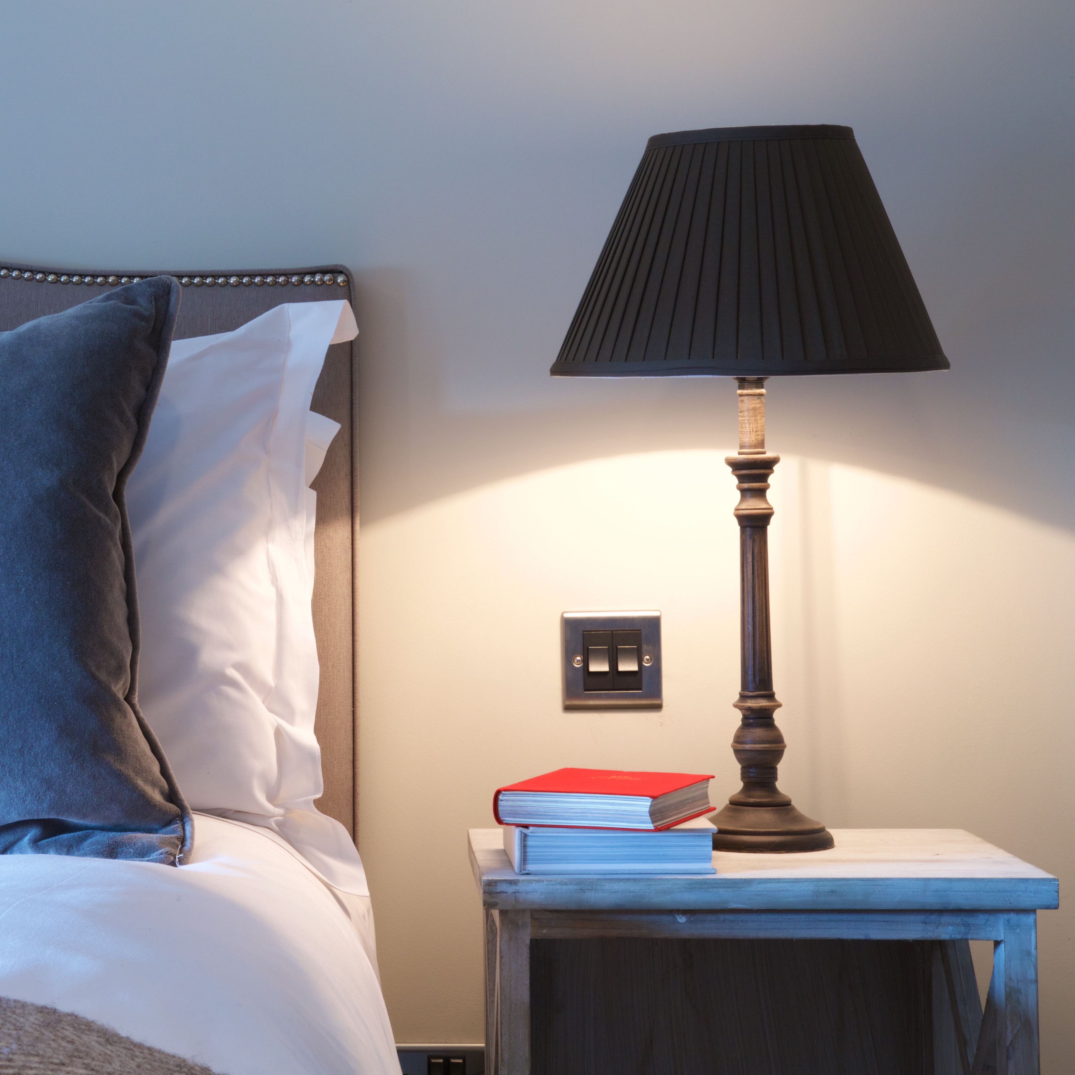 Una cama bien hecha con almohadas, una mesita de noche con lámpara y libros, y un interruptor de pared.