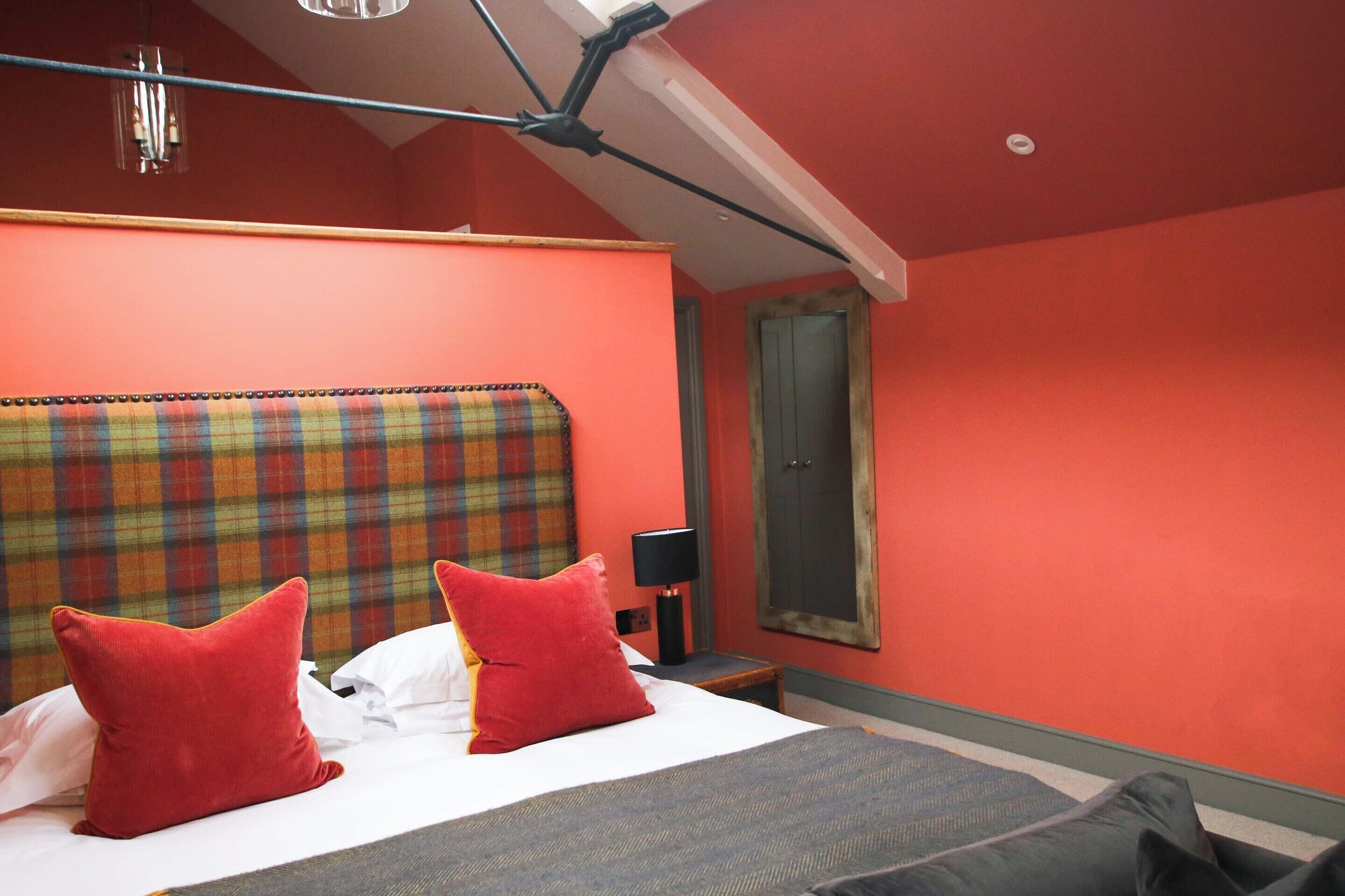 A hotel room with a plaid headboard, red pillows, a bed with a grey bedspread, a nightstand with a lamp, and a window with a curtain.