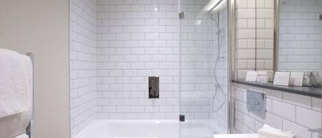 Un baño moderno con paredes de azulejos blancos, una ducha con cerramiento de vidrio, una bañera blanca y un lavabo con espejo.