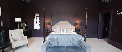 A bedroom with a four-poster bed, a light blue sofa, a white armchair, and two bedside tables with lamps.