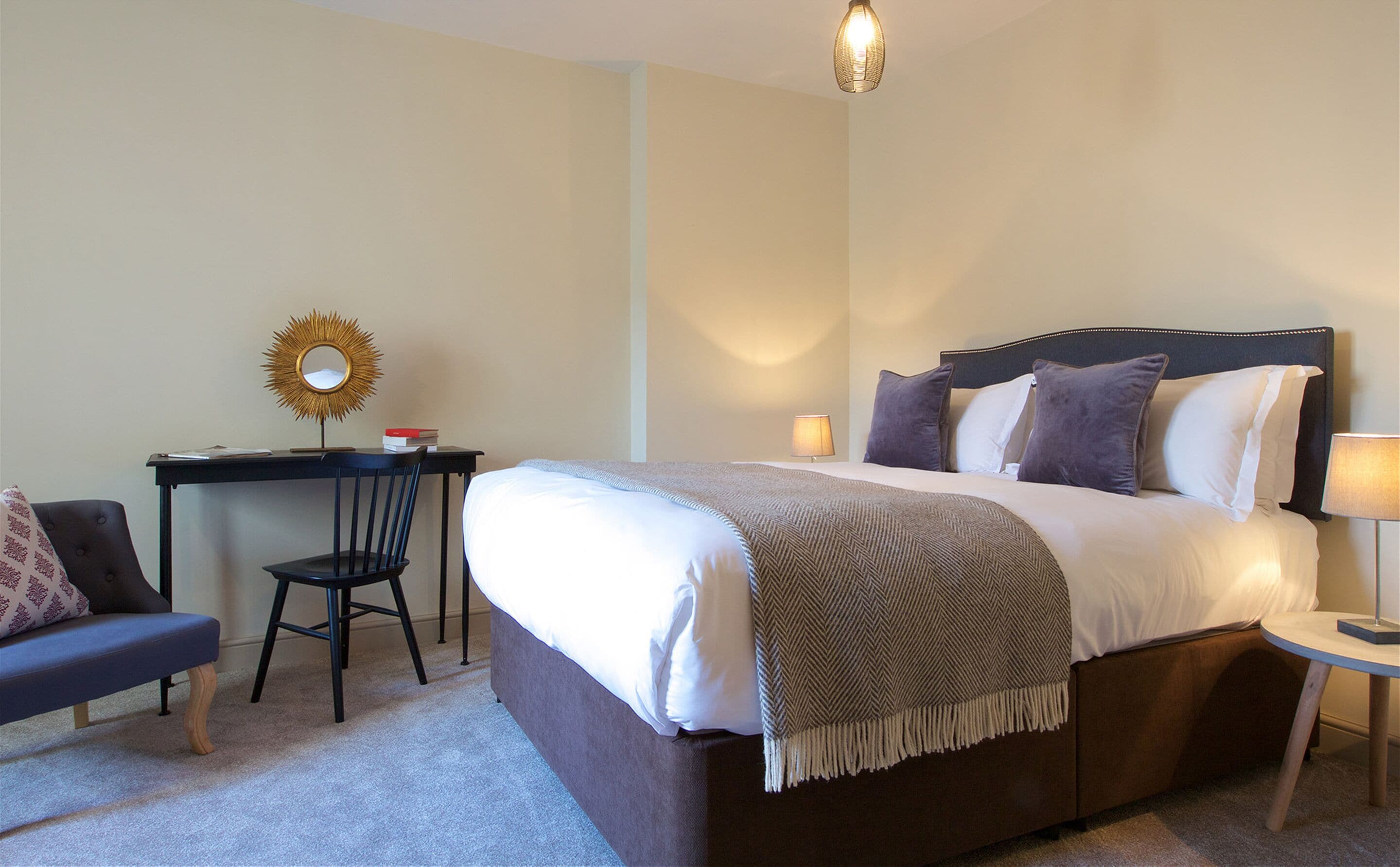 A neatly arranged hotel room with a large bed, a desk with a chair, a small table with a lamp, and a wall-mounted mirror.