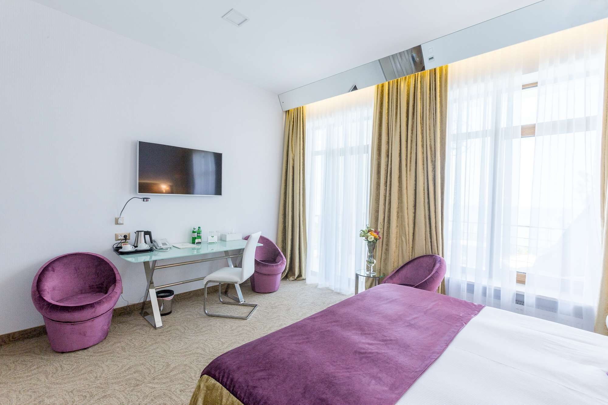 A hotel room with a bed, a desk with a chair, a TV mounted on the wall, and a window with curtains.