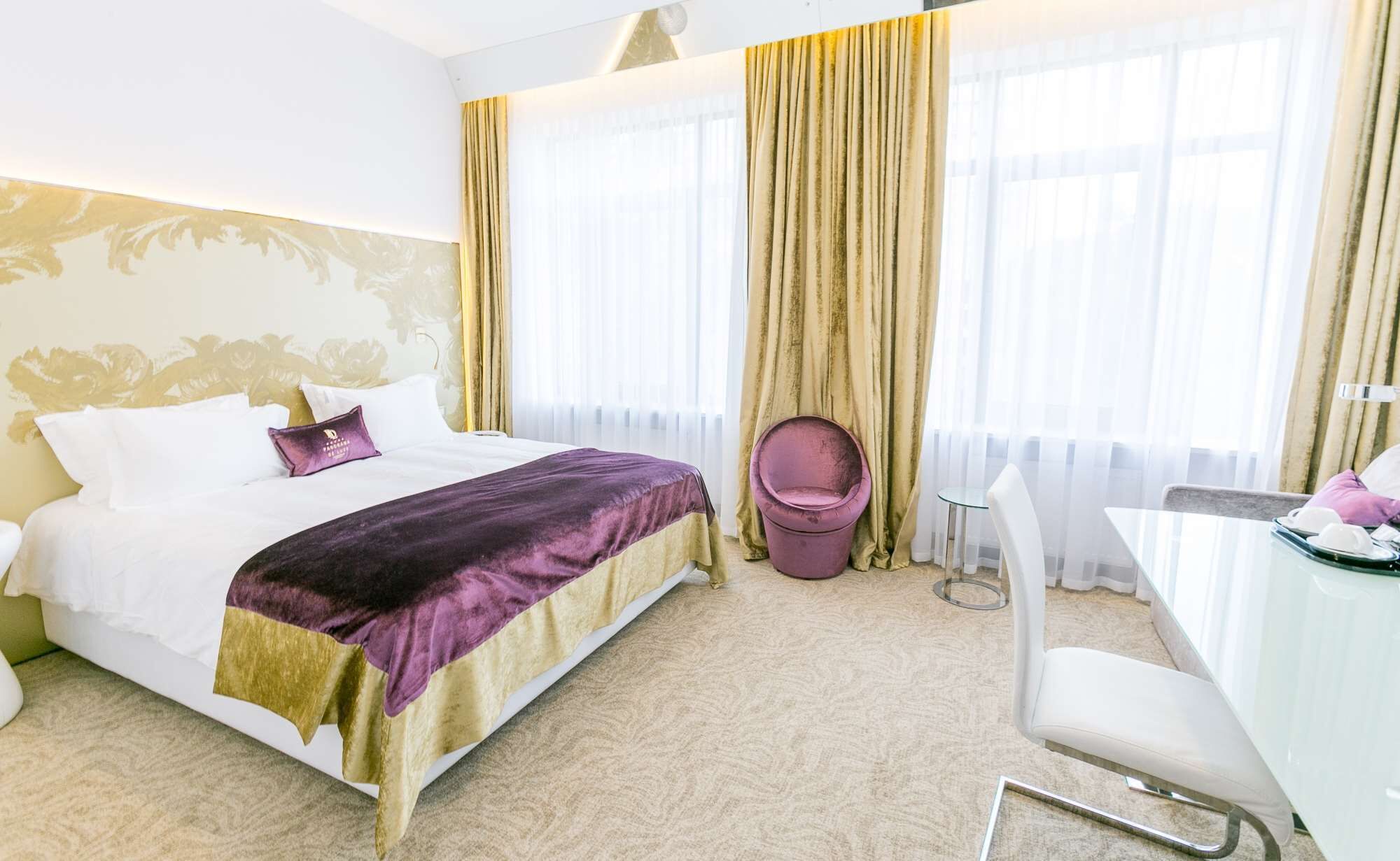 Une chambre d’hôtel avec un lit, un bureau, une chaise et de grandes fenêtres équipées de rideaux.