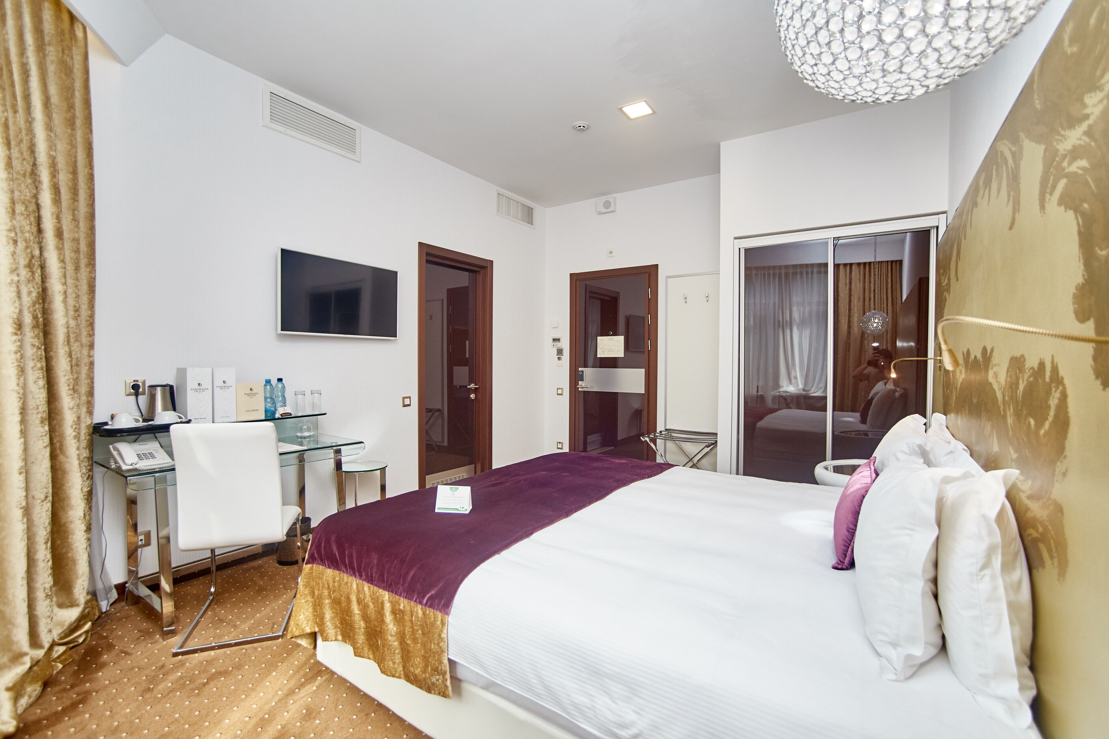 A hotel room with a bed, a desk with a chair, a TV, and a bathroom visible through an open door.