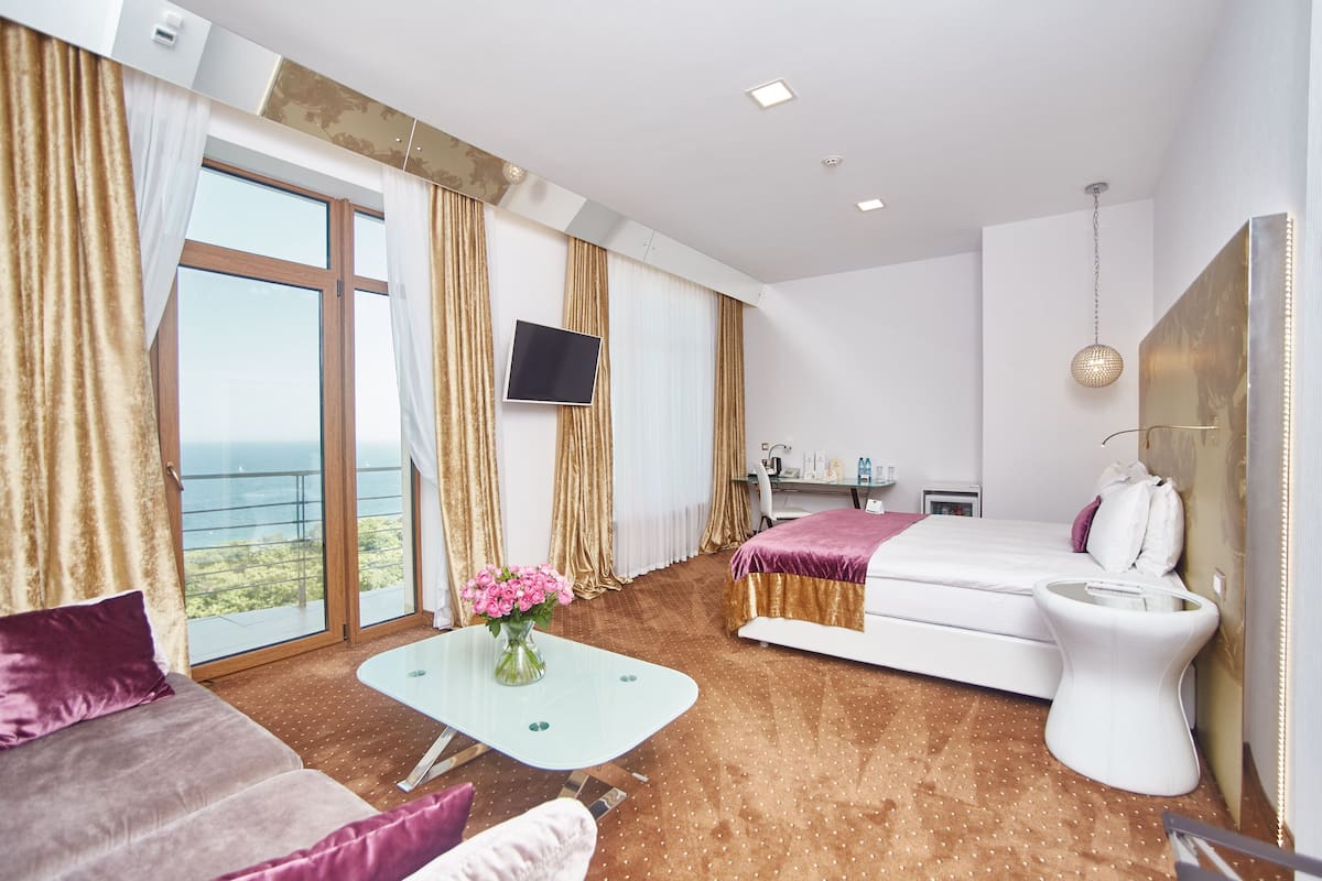 A modern hotel room with a large window, a bed, a sofa, a small table with a vase of flowers, and a TV on the wall.