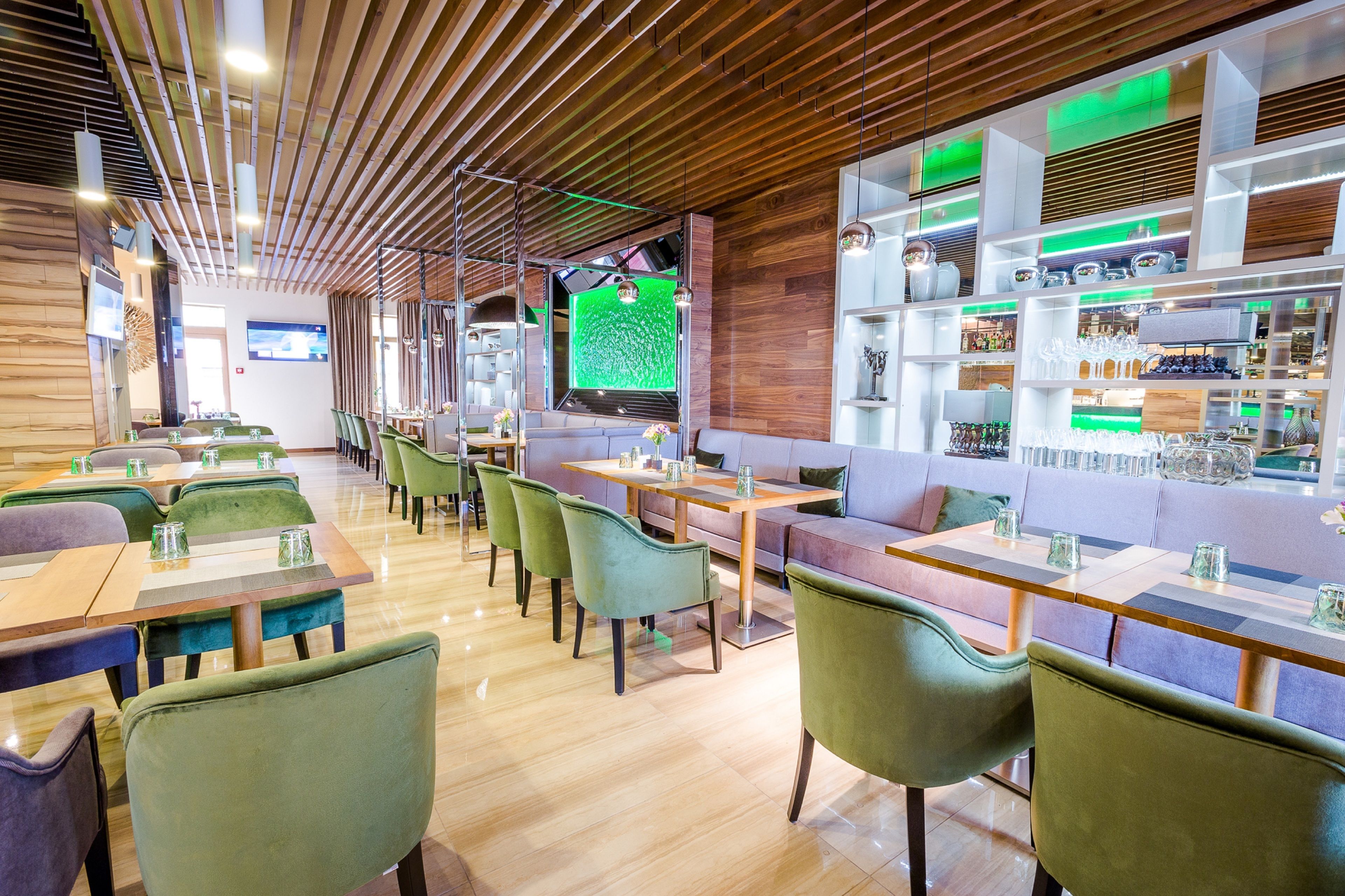A modern restaurant with wooden ceiling panels, green upholstered chairs, and a bar area with shelves of bottles and glasses.