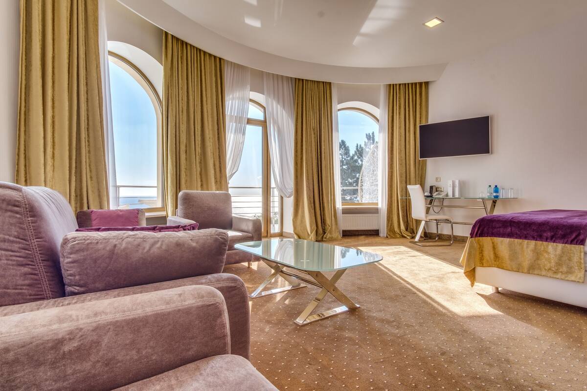 A modern hotel room with a sofa, a glass-top coffee table, a bed with a purple and gold bedspread, a flat-screen TV mounted on the wall, and large windows with curtains.