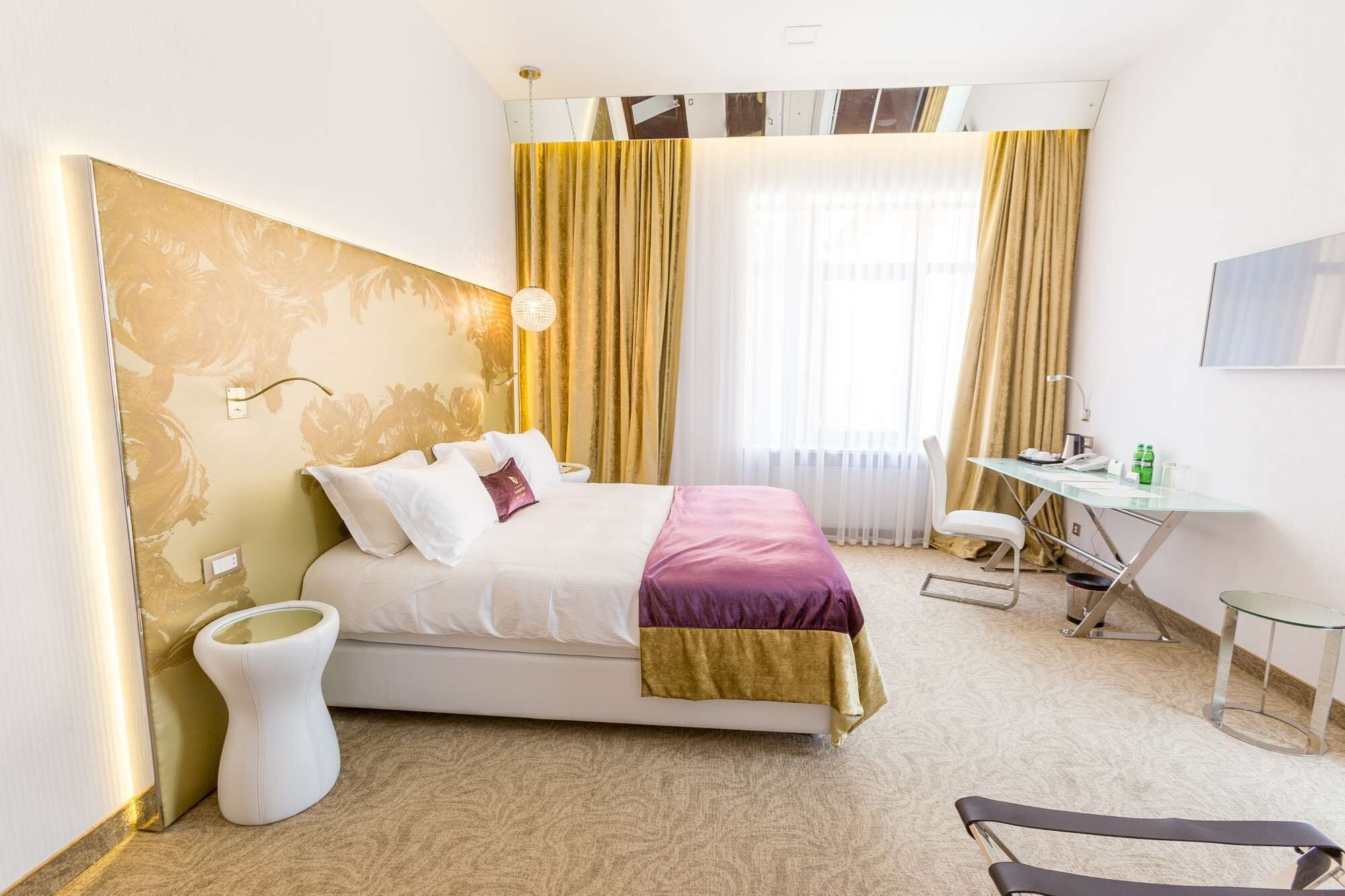 Habitación de hotel con cama, escritorio, silla, mesita de noche y un amplio ventanal con cortinas.