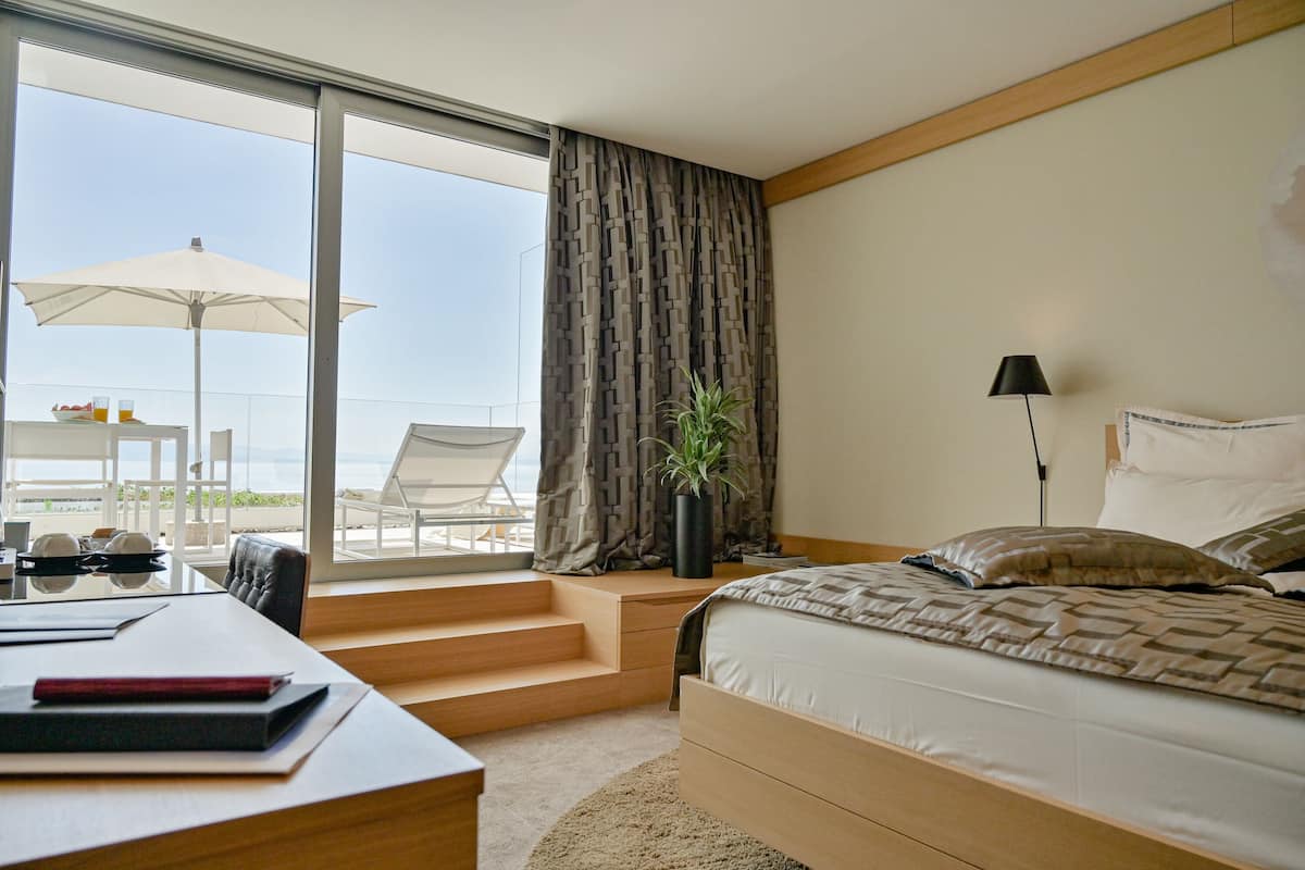 A modern hotel room with a large bed, a desk with a chair, a potted plant, and a view of the beach through large windows.