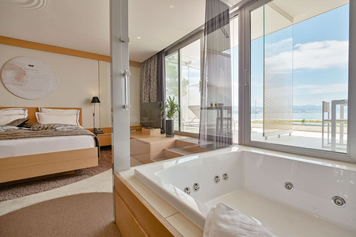 A modern bedroom with a large bed, a bathtub, and a balcony with a view.