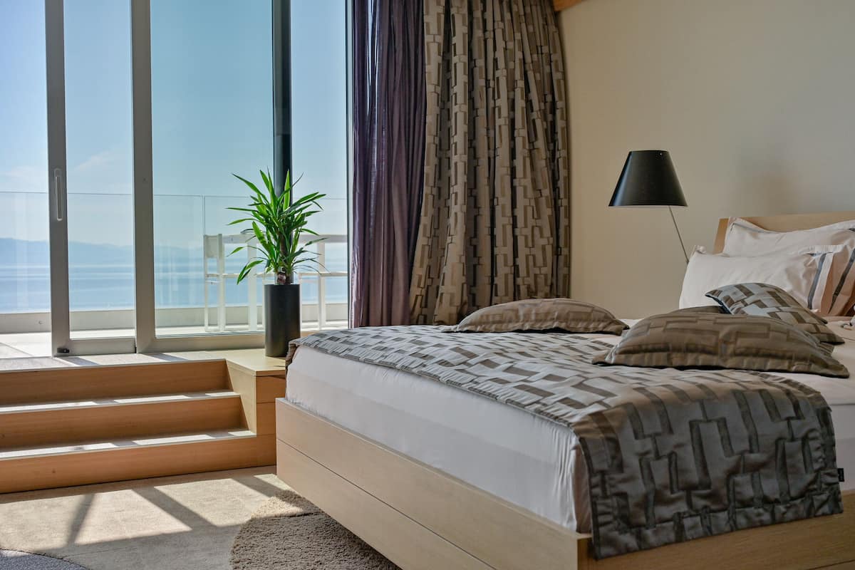 A bedroom with a large bed, a potted plant, a lamp, and a view of the sea through a sliding door.