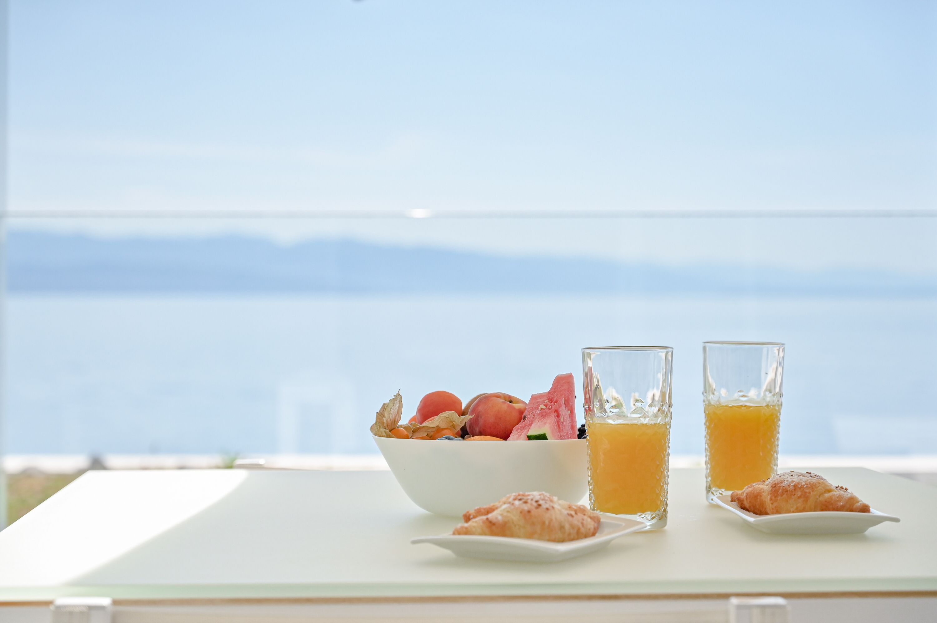 A breakfast setting with a bowl of fruit, two glasses of orange juice, and a croissant on a table with a view of the sea.