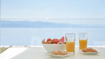 A breakfast setting with a bowl of fruit, two glasses of orange juice, and a croissant on a table with a view of the sea.