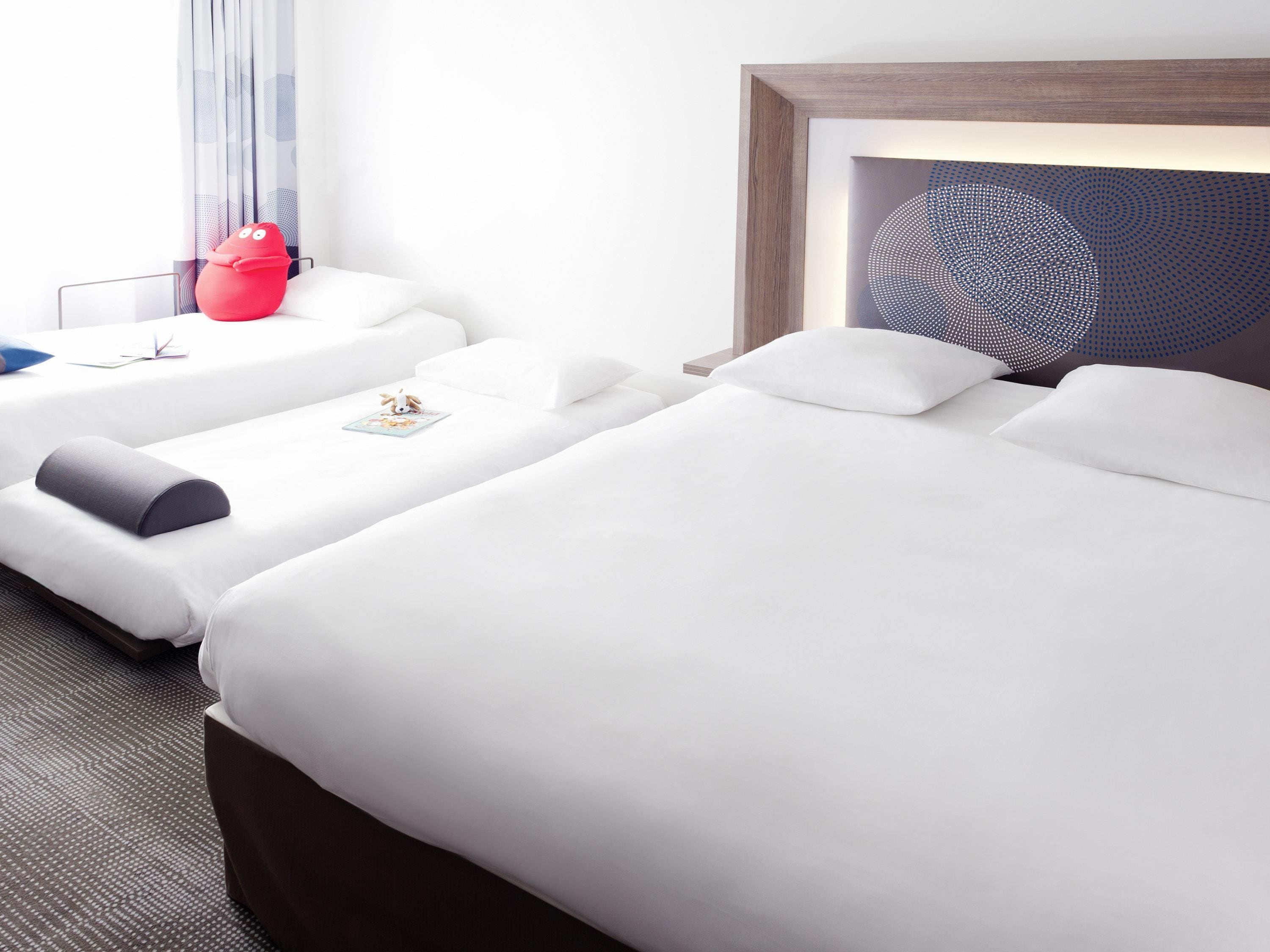 Habitación de hotel con dos camas, un juguete rojo, una obra de arte enmarcada y una ventana con cortinas.