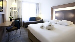 A hotel room with a bed, a desk, a chair, a lamp, and a window with curtains. - Novotel Basel City (Basel)