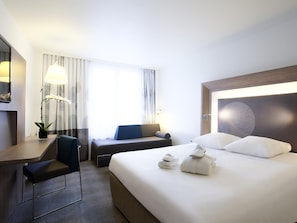A hotel room with a bed, a desk, a chair, a lamp, and a window with curtains. - Novotel Basel City (Basel)